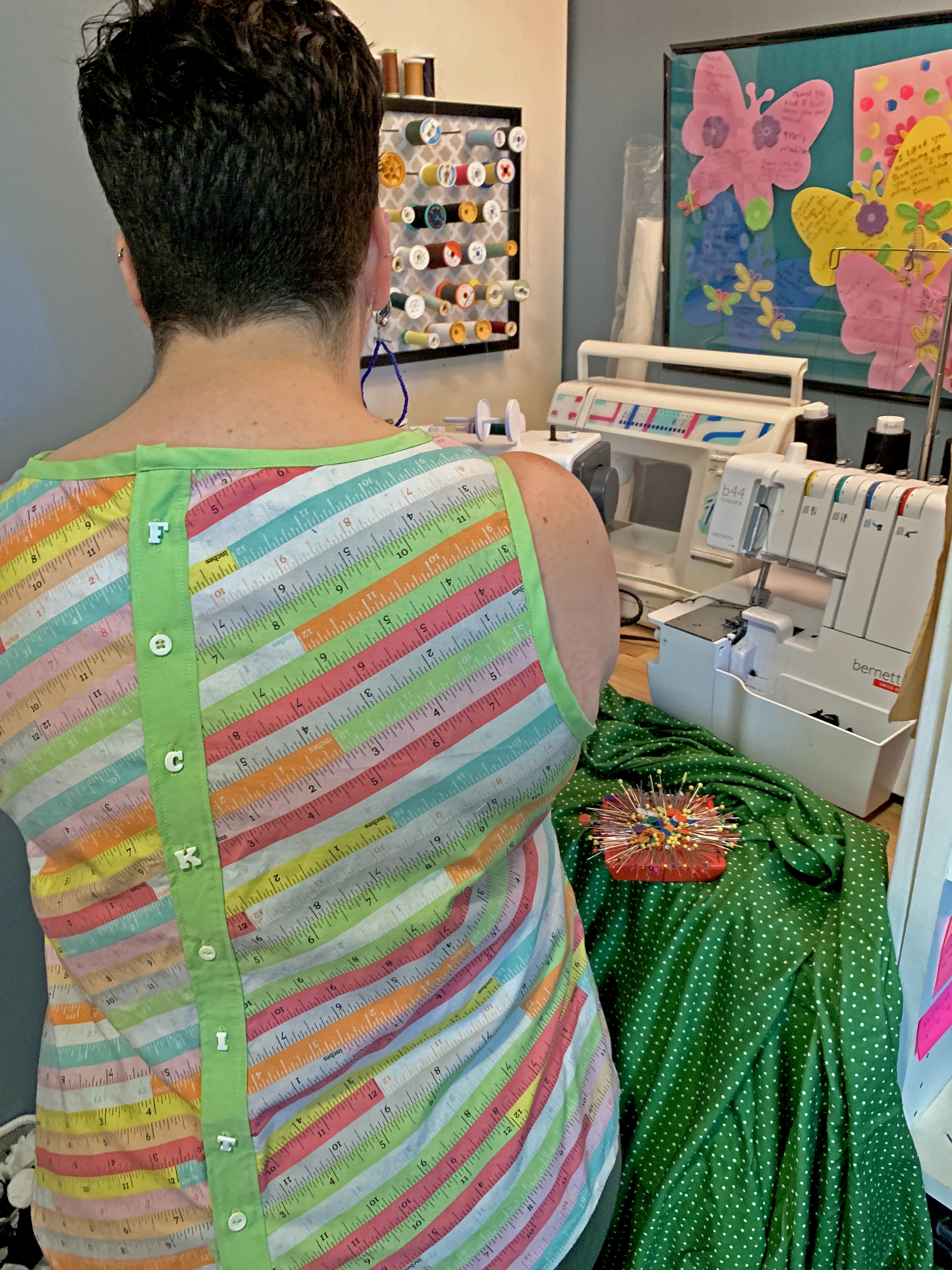 view over the shoulder of a woman with short hair using a sewing machine. There are letter shaped buttons down the back of her shirt spelling 'fuck it'