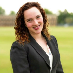 photo of a smiling white woman with shoulder length, curly, brown hair. Wearing a blazer