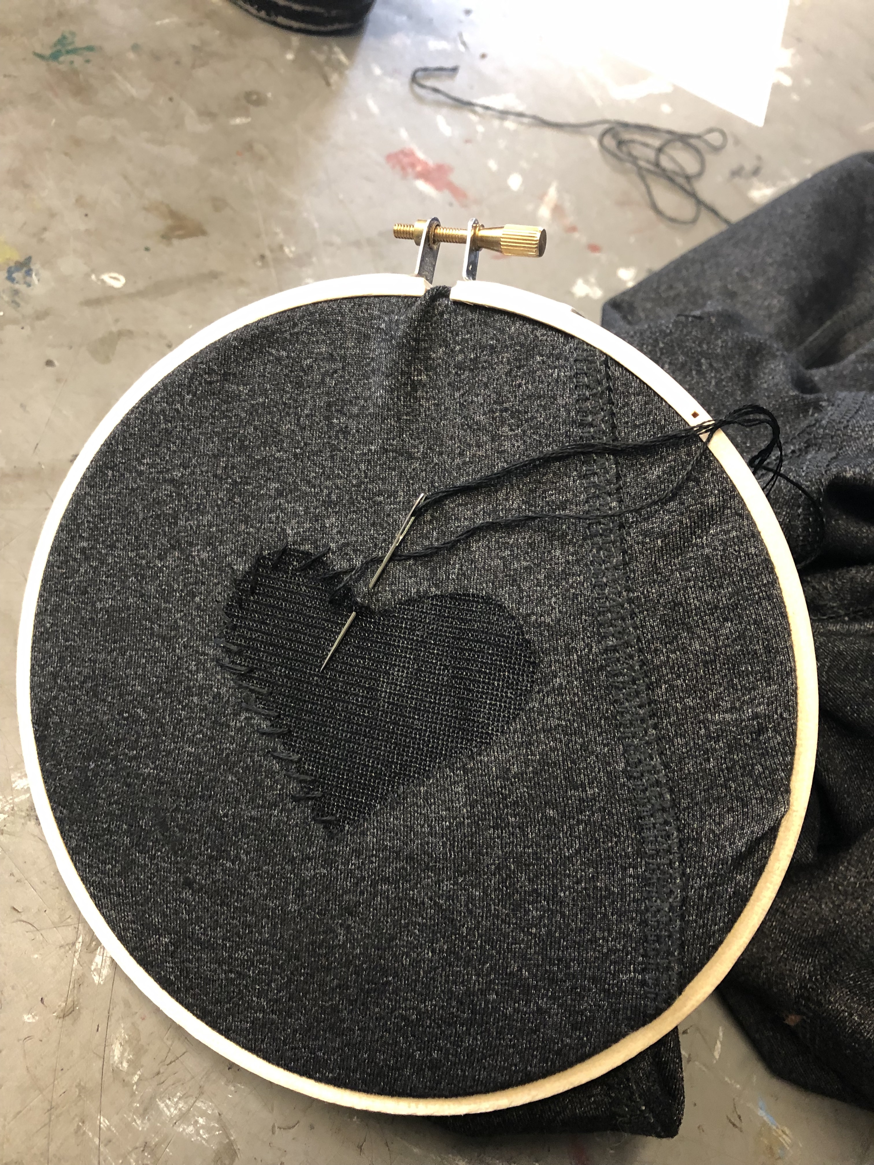 an embroidery hoop with a piece of gray fabric and a patch in the shape of a heart partially sewn on